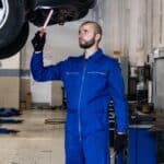 Automechanik v modré kombinéze prohlíží otevřenou kapotu osobního auta v dílně autoservisu