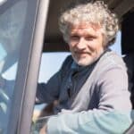 Starší muž se šedivými vlasy řídí automobil a drží volant oběma rukama