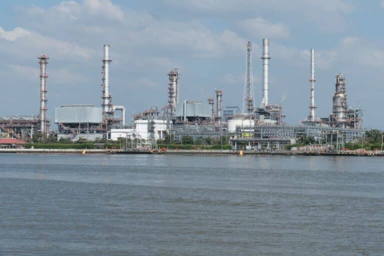 Rozlehlý komplex ropné rafinerie u vody pod zataženou oblohou.