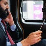 Muž v obleku sedí v autě, v jedné ruce drží mobilní telefon a na kolenou má tablet