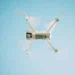 Bílý dron se čtyřmi rotory visí ve vzduchu na pozadí modré oblohy