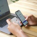 Člověk drží platební kartu a mobilní telefon u otevřeného notebooku