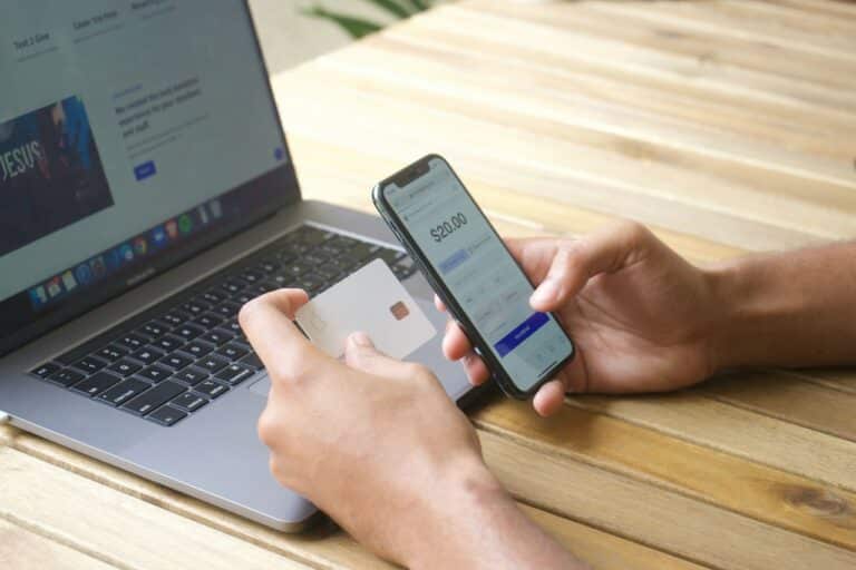 Člověk drží platební kartu a mobilní telefon u otevřeného notebooku