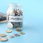 Skleněná dóza plná mincí s nálepkou Savings vedle kalkulačky na modrém podkladu