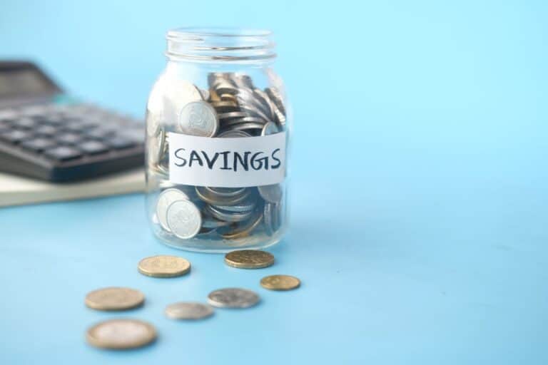 Skleněná dóza plná mincí s nálepkou Savings vedle kalkulačky na modrém podkladu
