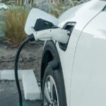 Bílý elektromobil stojí na parkovišti připojený kabelem k nabíjecí stanici