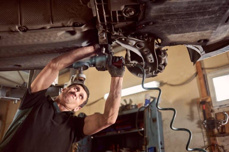 Automechanik v černém tričku utahuje utahovákem díly podvozku u kola zvednutého auta v dílně