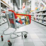 Nákupní vozík s červeným madlem plný potravin stojí v uličce supermarketu mezi regály
