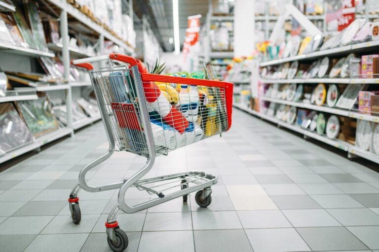 Nákupní vozík s červeným madlem plný potravin stojí v uličce supermarketu mezi regály